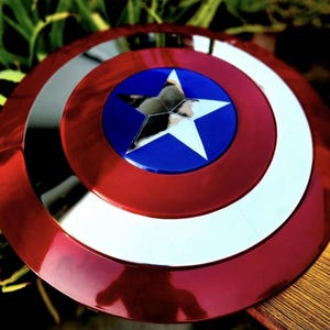 May include: A circular shield featuring a concentric design with a metallic red outer ring, a silver middle ring, and a central blue circle containing a large, five-pointed silver star. The surface has a reflective, polished finish throughout.