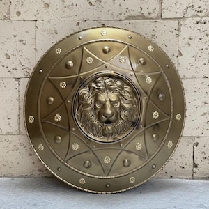 Puede incluir: Un escudo circular de latón que presenta un relieve tridimensional prominente de una cabeza de león en el centro. La superficie está decorada con un patrón repetitivo de pequeñas rosetas florales y tachuelas circulares en relieve.