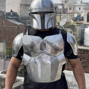 May include: A metallic silver Mandalorian-style helmet and chest armor set featuring a smooth, polished finish. The armor includes a full-face helmet with a T-shaped visor, a segmented chest plate, and matching shoulder pauldrons worn over a black shirt.