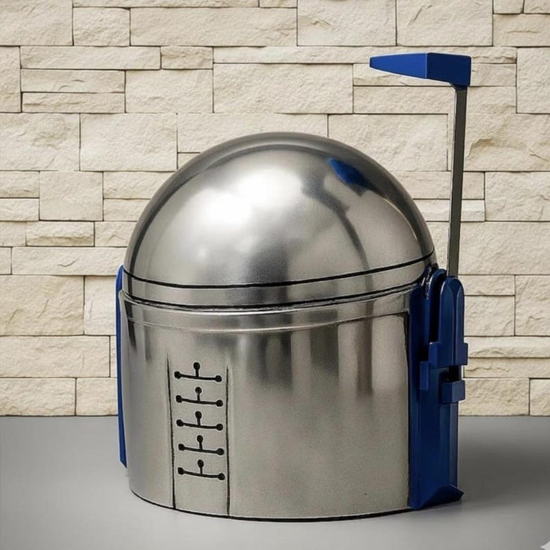 May include: A metallic silver helmet with a rounded dome top and a flat, vertical blue antenna extending from the right side. The front features a central vertical panel with five horizontal black line details, and blue accents on the sides.