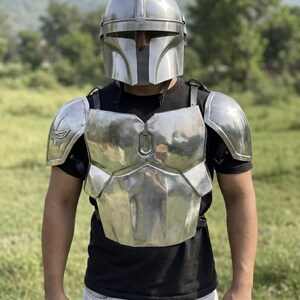 May include: A polished silver metal chest plate armor set with matching shoulder pauldrons and a full-face helmet featuring a T-shaped visor. The armor is worn over a black short-sleeved t-shirt and light-colored pants, showcasing a reflective finish.