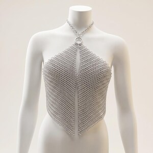 May include: A silver-colored chainmail halter top constructed from interlocking metal rings. The garment features a central circular ring at the neckline that connects to a chain choker, with two pointed panels draping down the torso.