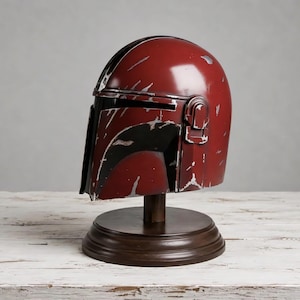 May include: A dark red helmet with a black T-shaped visor and silver-colored weathering marks across the surface. The helmet is mounted on a circular dark brown wooden stand, featuring a smooth dome and a distinct side ear piece detail.