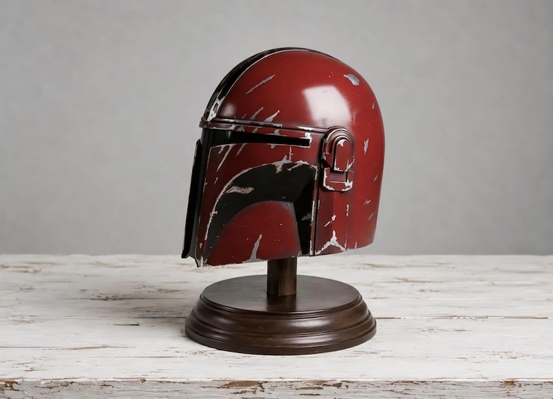 May include: A dark red helmet with a black T-shaped visor and silver-colored weathering marks across the surface. The helmet is mounted on a circular dark brown wooden stand, featuring a smooth dome and a distinct side ear piece detail.