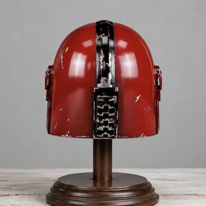 May include: A red helmet with a distressed finish and a black vertical stripe running down the center. The helmet features a rectangular detail on the back and is mounted on a dark brown circular wooden stand with a central pedestal base.