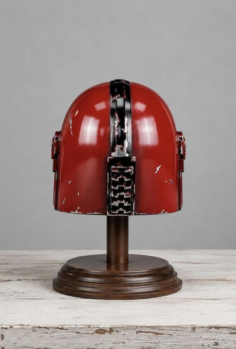 May include: A red helmet with a distressed finish and a black vertical stripe running down the center. The helmet features a rectangular detail on the back and is mounted on a dark brown circular wooden stand with a central pedestal base.