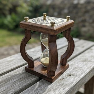 May include: A wooden hourglass featuring a dark brown frame with four curved supports. The top platform is a square brass plate with a black and white checkered pattern and four small brass knobs at the corners. Clear glass bulbs contain sand.