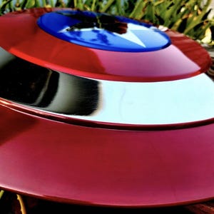 May include: A circular shield featuring a glossy red outer ring, a polished silver middle ring, and a vibrant blue center circle. A large, white five-pointed star is centered within the blue circle, creating a distinct, high-contrast design.