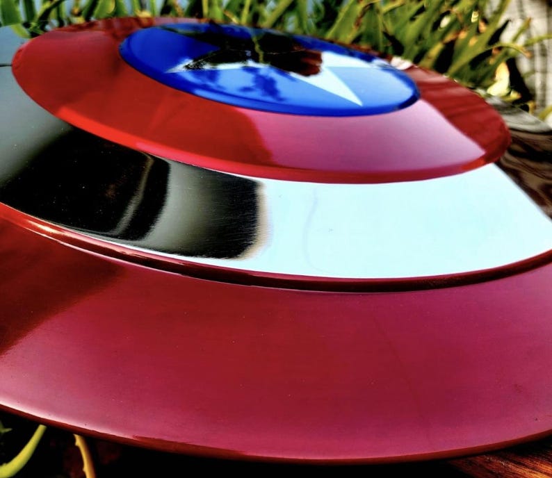May include: A circular shield featuring a glossy red outer ring, a polished silver middle ring, and a vibrant blue center circle. A large, white five-pointed star is centered within the blue circle, creating a distinct, high-contrast design.