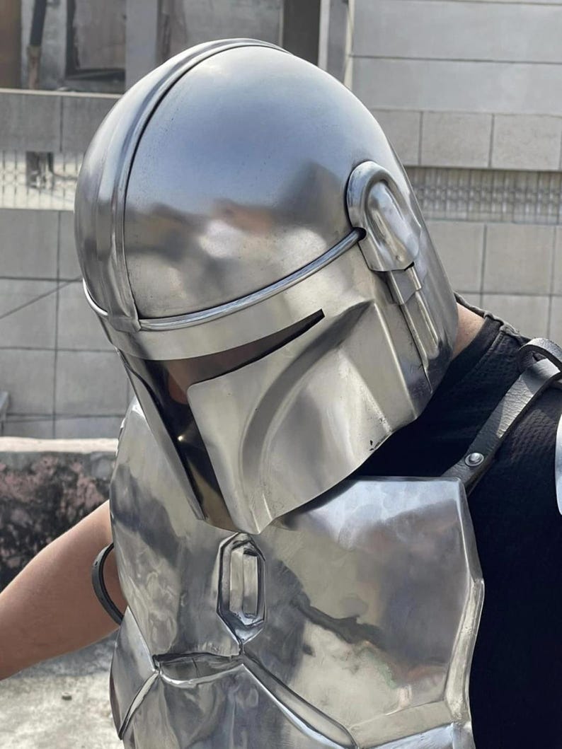 May include: A polished silver-colored metal helmet with a T-shaped visor opening and a matching chest plate. The helmet features a smooth, rounded dome and integrated side ear pieces, while the chest armor includes a central vertical panel.