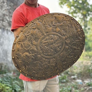 Pode incluir: Um escudo grande de formato oval na cor bronze com relevos intrincados de figuras mitológicas, folhagens e animais. A borda é decorada com uma série de tachas metálicas espaçadas uniformemente ao longo de todo o contorno externo.