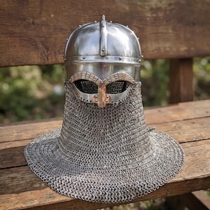 May include: A polished steel helmet featuring a rounded dome, a central vertical ridge, and a protective eye guard with decorative brass-toned accents. A dense, silver-colored chainmail aventail hangs from the base, covering the neck and shoulders.