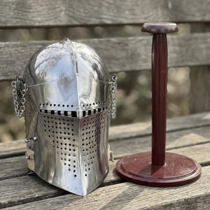 Op de afbeelding: Een gepolijste zilverkleurige metalen ridderhelm met een afgerond vizier met een horizontale oogopening en een geperforeerde voorplaat, vergezeld van een donkerbruine houten standaard met een ronde voet en een verticale paal.