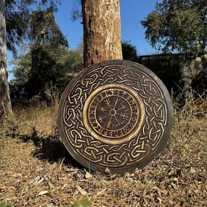 Op de afbeelding: Een ronde houten wandplaat met een donkerbruine afwerking en ingewikkelde uitgesneden Keltische knoopmotieven rondom een centraal Vegvisir-symbool. Het ontwerp bevat een ring van runentekens die het centrale kompasembleem omcirkelt.