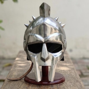 Könnte beinhalten: Ein polierter, silberfarbener Metallhelm mit einem markanten Mittelkamm, einer stilisierten Gesichtsmaske mit breiten Augenöffnungen und einer Reihe kleiner, spitzer Dornen entlang der oberen Kuppel. Ein dünner brauner Lederriemen hängt seitlich.