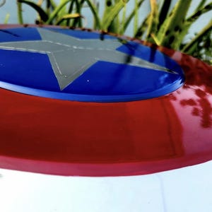 May include: A circular shield featuring a glossy red outer ring and a vibrant blue center circle. A large, five-pointed silver star is centered within the blue circle. The surface has a smooth, reflective finish with visible light highlights.