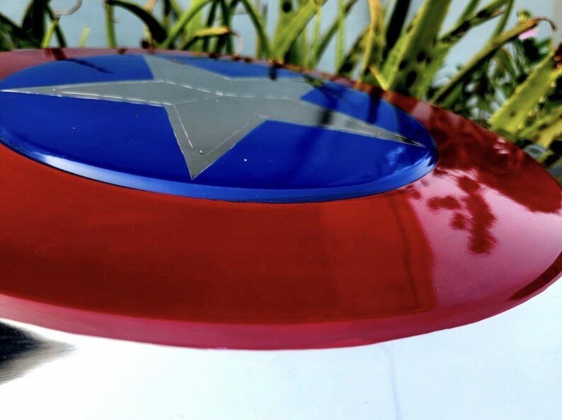 May include: A circular shield featuring a glossy red outer ring and a vibrant blue center circle. A large, five-pointed silver star is centered within the blue circle. The surface has a smooth, reflective finish with visible light highlights.