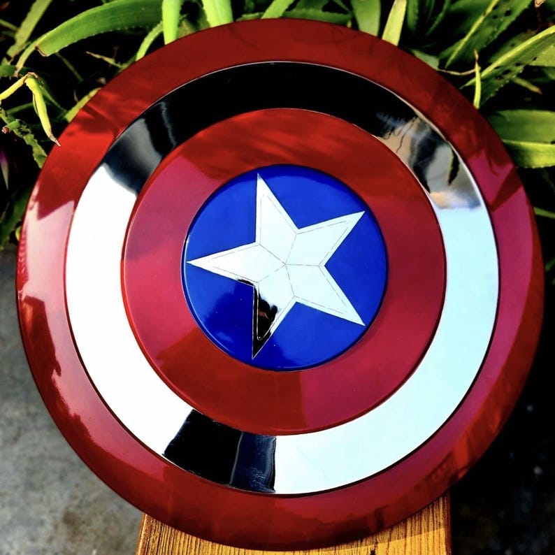 May include: A circular shield featuring a glossy red outer ring, a reflective silver middle ring, and a deep blue center circle. A large, five-pointed white star is centered within the blue circle, creating a bold, high-contrast design.