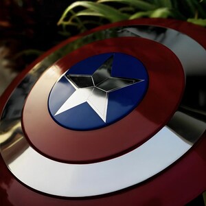 May include: A circular shield featuring a metallic silver star centered on a blue circular background, surrounded by a wide silver ring and an outer deep red border. The surface has a reflective, polished finish with concentric circular sections.