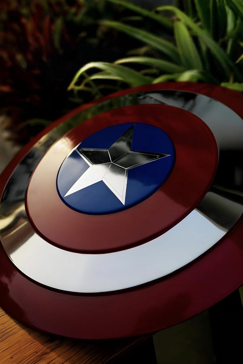 May include: A circular shield featuring a metallic silver star centered on a blue circular background, surrounded by a wide silver ring and an outer deep red border. The surface has a reflective, polished finish with concentric circular sections.