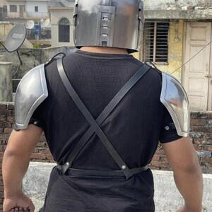May include: A metallic silver helmet with a smooth, rounded dome and a vertical rectangular vent design on the back, paired with matching silver shoulder pauldrons secured by black leather straps that cross over the back of a black t-shirt.
