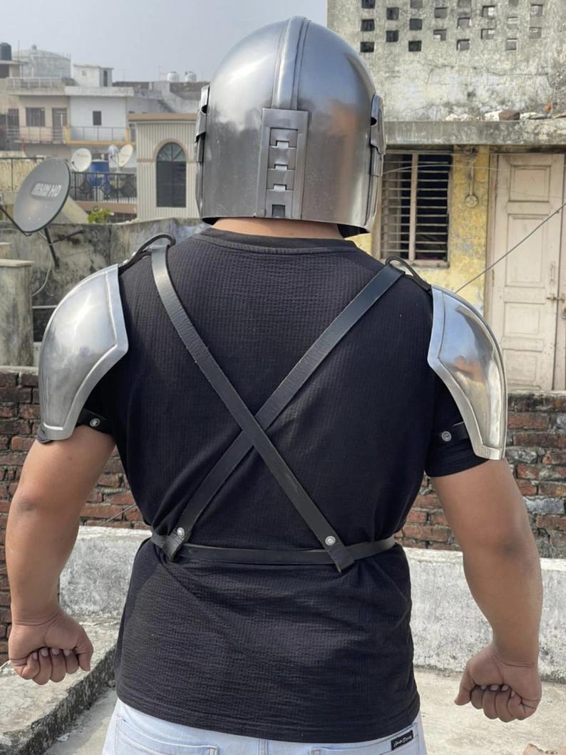May include: A metallic silver helmet with a smooth, rounded dome and a vertical rectangular vent design on the back, paired with matching silver shoulder pauldrons secured by black leather straps that cross over the back of a black t-shirt.
