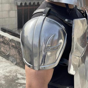 May include: A polished silver metal shoulder pauldron featuring a raised crescent-shaped emblem, secured with black leather straps over a black shirt. The armor piece is part of a larger set including a matching chest plate and a metallic helmet.