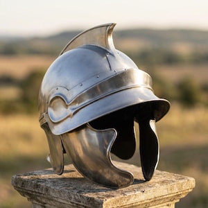 May include: A polished silver-colored metal helmet featuring a prominent central crest, a curved visor, and side cheek guards. The helmet is constructed with visible rivets along the brow and side panels, showcasing a smooth, reflective finish.