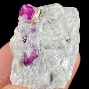 May include: A raw mineral specimen featuring a large, vibrant magenta-pink crystal embedded within a textured, translucent white and light grey matrix. The stone displays irregular, jagged edges and a crystalline surface structure throughout.