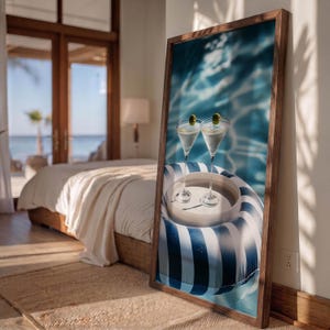 May include: A rectangular framed wall art piece featuring a photograph of two martini glasses with green olives resting on a white circular tray, which is placed inside a blue and white striped inflatable pool float on shimmering blue water.