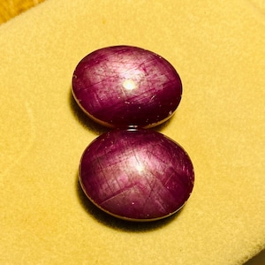 Könnte beinhalten: Zwei ovale, tief magentafarbene Cabochon-Edelsteine mit einer glatten, polierten Oberfläche. Die Steine weisen eine natürliche, faserige Textur auf, die unter der Oberfläche sichtbar ist und das Licht mit einem subtilen Glanz reflektiert.