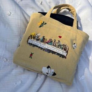 May include: A corduroy tote bag in a light yellow color, featuring an embroidered depiction of the Last Supper. The bag has a black interior and is adorned with floral and heart-shaped embellishments. The bag has a handle.