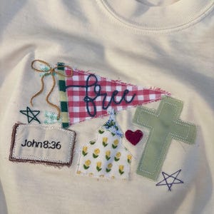 May include: A cream-colored sweatshirt featuring an embroidered applique design. The design includes a red and white checkered pennant with the word free, a book labeled John 8:36, a floral church, a red heart, a green cross, and two stars.