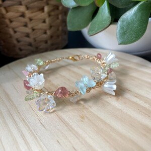 Pode incluir: Uma pulseira de fio em tom dourado com um sortido de contas em forma de flor transl&uacute;cidas e opacas em branco, azul claro e transparente, intercaladas com pequenas contas em forma de folha verde e detalhes em tons de vermelho rosado.