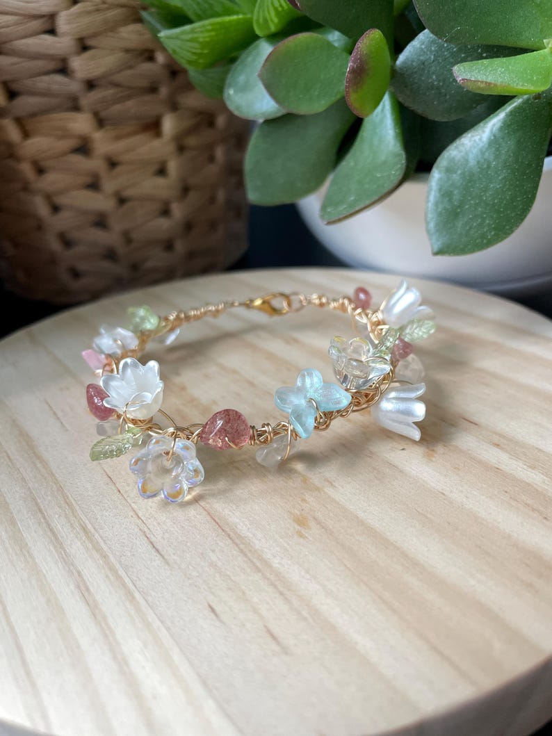 Pode incluir: Uma pulseira de fio em tom dourado com um sortido de contas em forma de flor transl&uacute;cidas e opacas em branco, azul claro e transparente, intercaladas com pequenas contas em forma de folha verde e detalhes em tons de vermelho rosado.