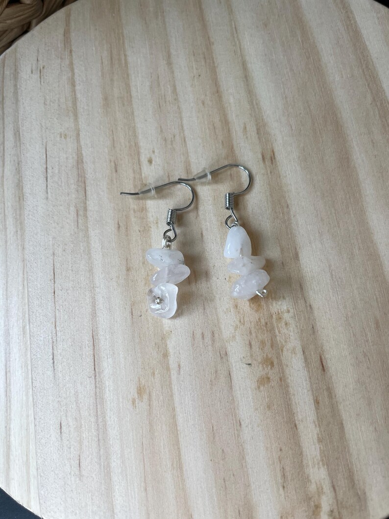 Puede incluir: Un par de pendientes colgantes con ganchos de metal plateado y peque&ntilde;as piedras blancas transl&uacute;cidas de forma irregular. Cada pendiente consta de tres piezas de piedra pulida apiladas suspendidas de un gancho de alambre sencillo.