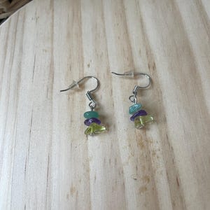 Puede incluir: Un par de pendientes colgantes con ganchos de metal plateado y peque&ntilde;as cuentas irregulares en forma de astilla en tonos verde claro, morado y azul verdoso. Cada pendiente consta de tres astillas de piedra apiladas en un gancho de metal.