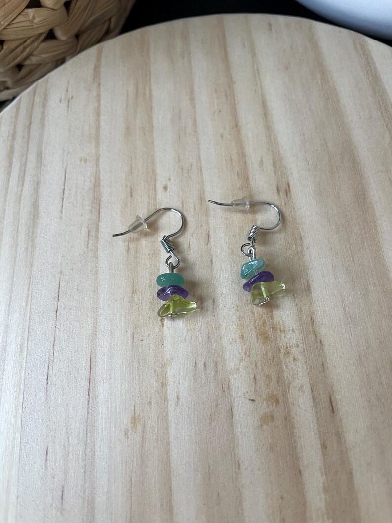 Puede incluir: Un par de pendientes colgantes con ganchos de metal plateado y peque&ntilde;as cuentas irregulares en forma de astilla en tonos verde claro, morado y azul verdoso. Cada pendiente consta de tres astillas de piedra apiladas en un gancho de metal.