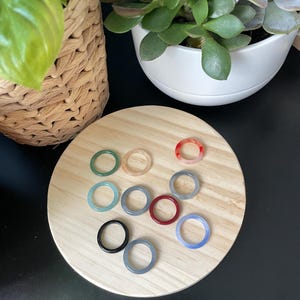 Könnte beinhalten: Eine Sammlung von zehn kreisförmigen Ringen in verschiedenen Farben, darunter Schwarz, Dunkelgrau, Hellgrau, Rot, Dunkelrot, Hellblau, Blaugrün und durchscheinendes Pfirsich, angeordnet auf einer runden, hellen Holzoberfläche mit sichtbarer Maserung.
