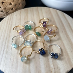 Puede incluir: Una colección de nueve anillos envueltos en alambre con pequeñas piedras preciosas pulidas en varios colores, incluyendo verde, morado, azul claro y azul oscuro. Los anillos están hechos de alambre de metal en tonos oro y plata.