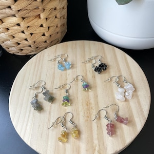 Puede incluir: Una colecci&oacute;n de siete pares de pendientes colgantes con peque&ntilde;as piedras preciosas pulidas e irregulares en varios colores, incluyendo azul, negro, verde, morado, amarillo, rosa y blanco, unidos a ganchos de metal de tono plateado.