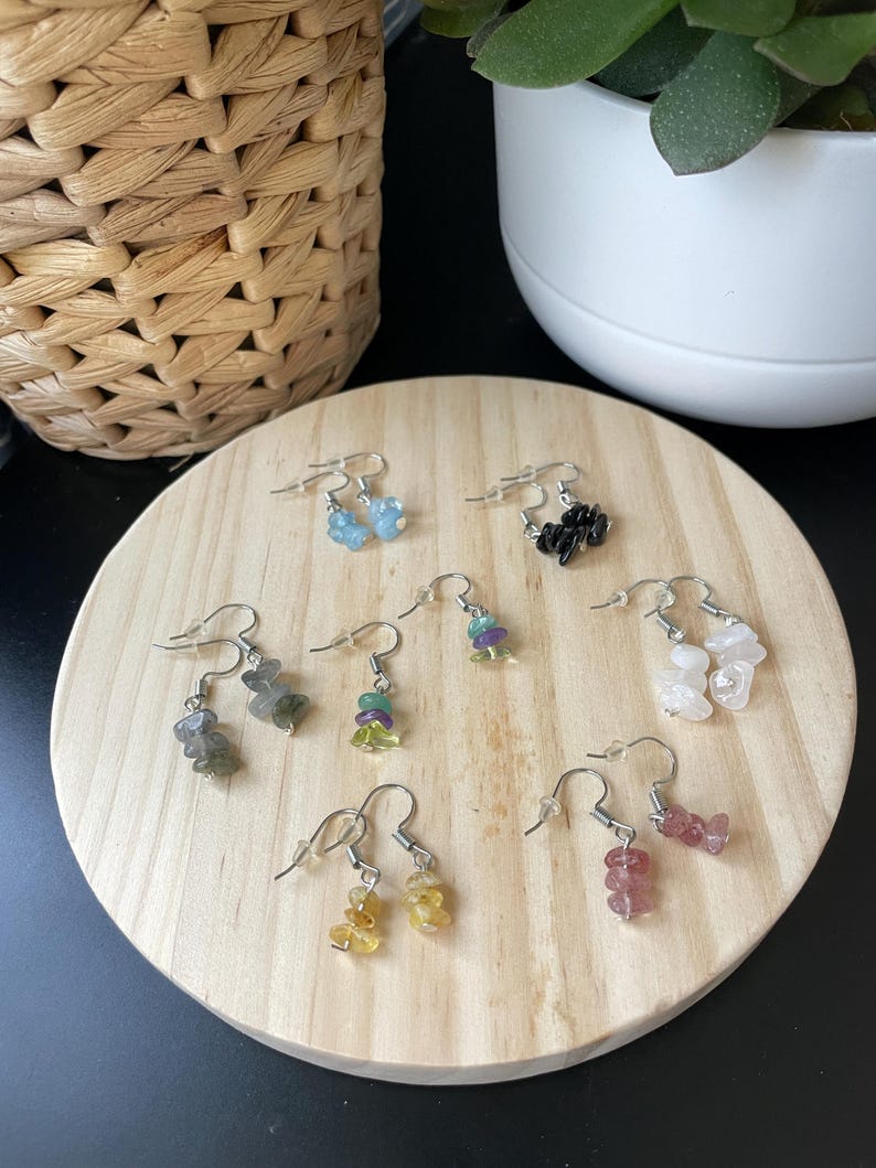 Puede incluir: Una colecci&oacute;n de siete pares de pendientes colgantes con peque&ntilde;as piedras preciosas pulidas e irregulares en varios colores, incluyendo azul, negro, verde, morado, amarillo, rosa y blanco, unidos a ganchos de metal de tono plateado.