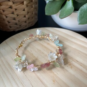 Pode incluir: Uma pulseira delicada feita de fio dourado, apresentando pequenas contas transl&uacute;cidas em forma de flor branca, detalhes em forma de folha verde-clara e fragmentos de pedra irregulares em rosa e azul-claro ao longo do aro.