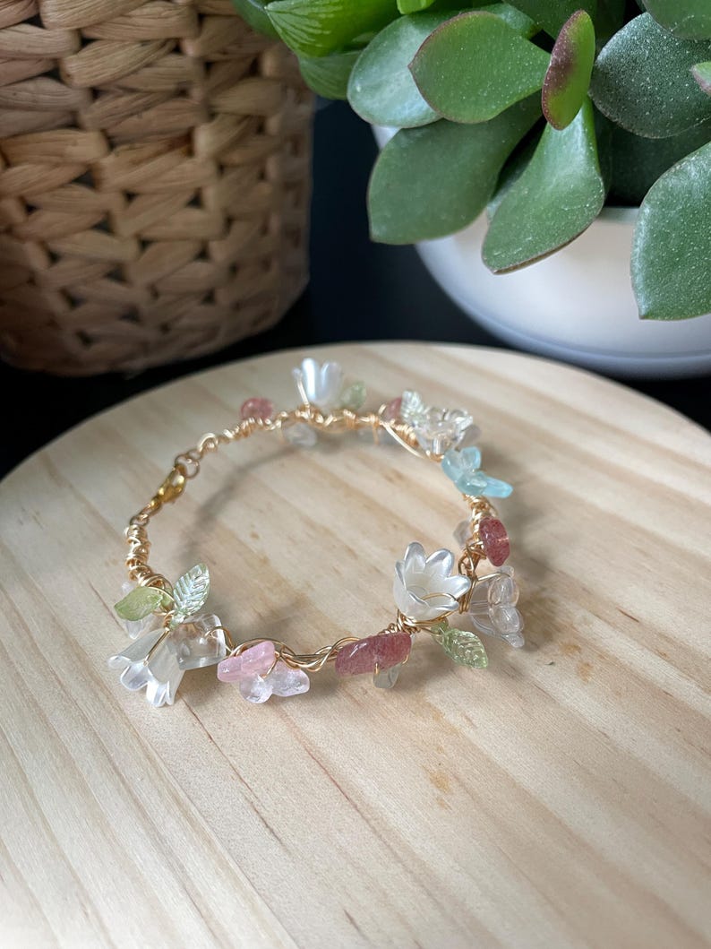 Pode incluir: Uma pulseira delicada feita de fio dourado, apresentando pequenas contas transl&uacute;cidas em forma de flor branca, detalhes em forma de folha verde-clara e fragmentos de pedra irregulares em rosa e azul-claro ao longo do aro.