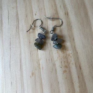 Puede incluir: Un par de pendientes colgantes con ganchos de metal plateado y cierres de pl&aacute;stico transparente. Cada pendiente consta de tres peque&ntilde;as piedras irregulares pulidas en tonos gris, gris azulado y verde apagado, suspendidas verticalmente.