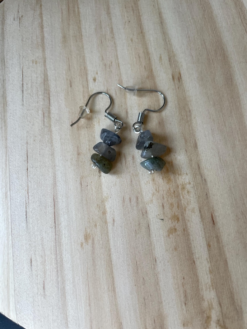 Puede incluir: Un par de pendientes colgantes con ganchos de metal plateado y cierres de pl&aacute;stico transparente. Cada pendiente consta de tres peque&ntilde;as piedras irregulares pulidas en tonos gris, gris azulado y verde apagado, suspendidas verticalmente.