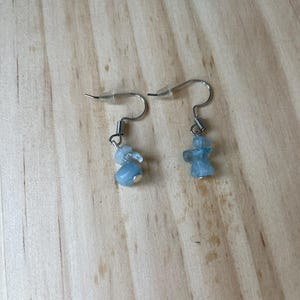 Puede incluir: Un par de pendientes colgantes con peque&ntilde;as piedras pulidas de color azul claro suspendidas de ganchos de metal plateado. Cada pendiente incluye un detalle de resorte en espiral sobre el grupo de piedras y un tope de pl&aacute;stico transparente.