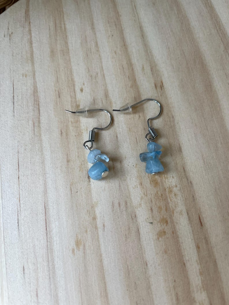 Puede incluir: Un par de pendientes colgantes con peque&ntilde;as piedras pulidas de color azul claro suspendidas de ganchos de metal plateado. Cada pendiente incluye un detalle de resorte en espiral sobre el grupo de piedras y un tope de pl&aacute;stico transparente.