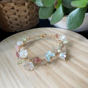 Pode incluir: Uma pulseira delicada feita de fio dourado, apresentando um arranjo de pequenas contas transl&uacute;cidas em forma de flor em tons de branco, azul claro e transparente, intercaladas com pequenos detalhes de pedra polida em rosa e verde.