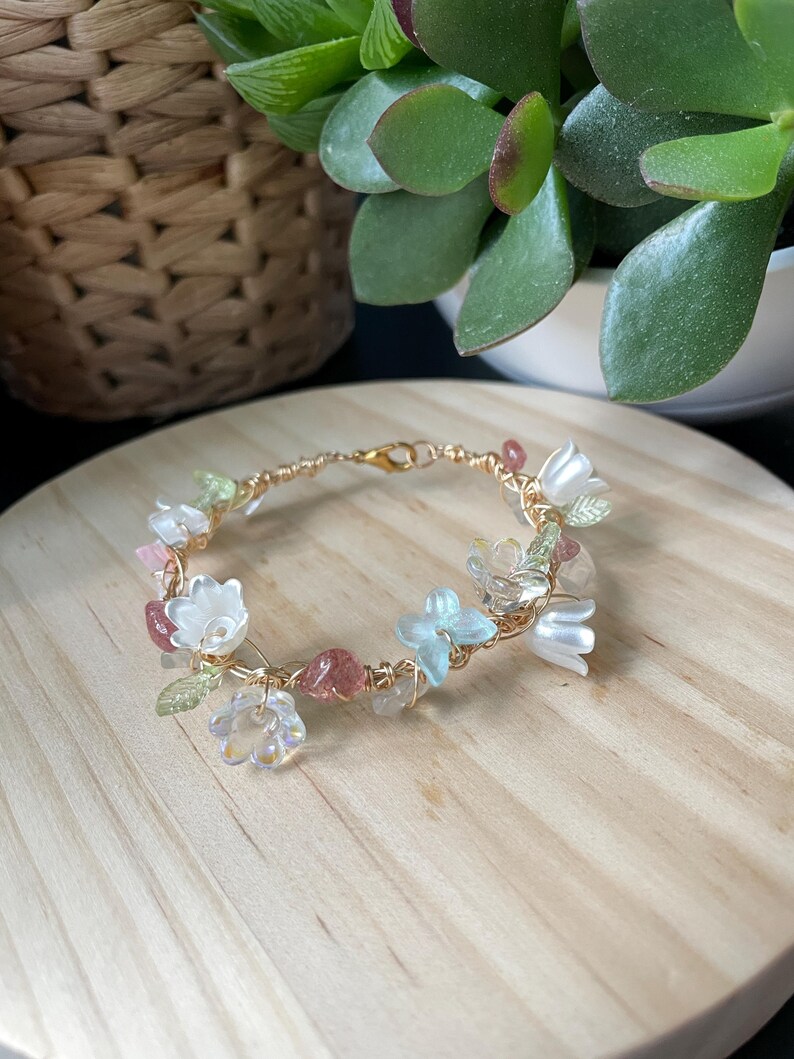 Pode incluir: Uma pulseira delicada feita de fio dourado, apresentando um arranjo de pequenas contas transl&uacute;cidas em forma de flor em tons de branco, azul claro e transparente, intercaladas com pequenos detalhes de pedra polida em rosa e verde.